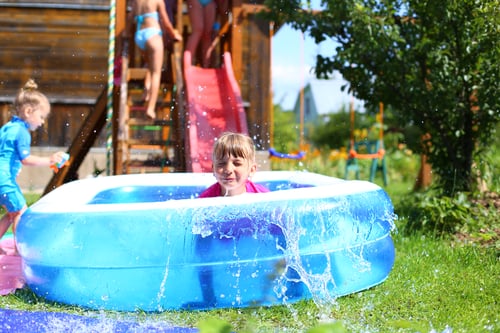 Little girl in swimsuit rolls down slide into inflatable rubber pool. Swim activity in backyard of country house.